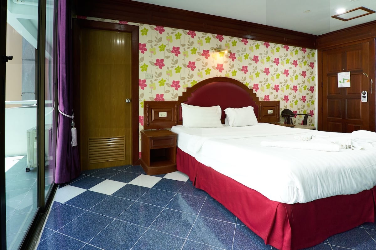 Una habitación de hotel con piso a cuadros, una cama con sábanas blancas, cabecera de madera y papel tapiz floral.