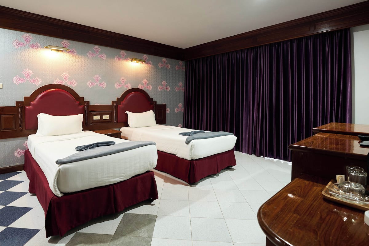 Habitación de hotel con dos camas, un cabecero de madera, suelo a cuadros, un escritorio con bandeja y una ventana con cortinas.