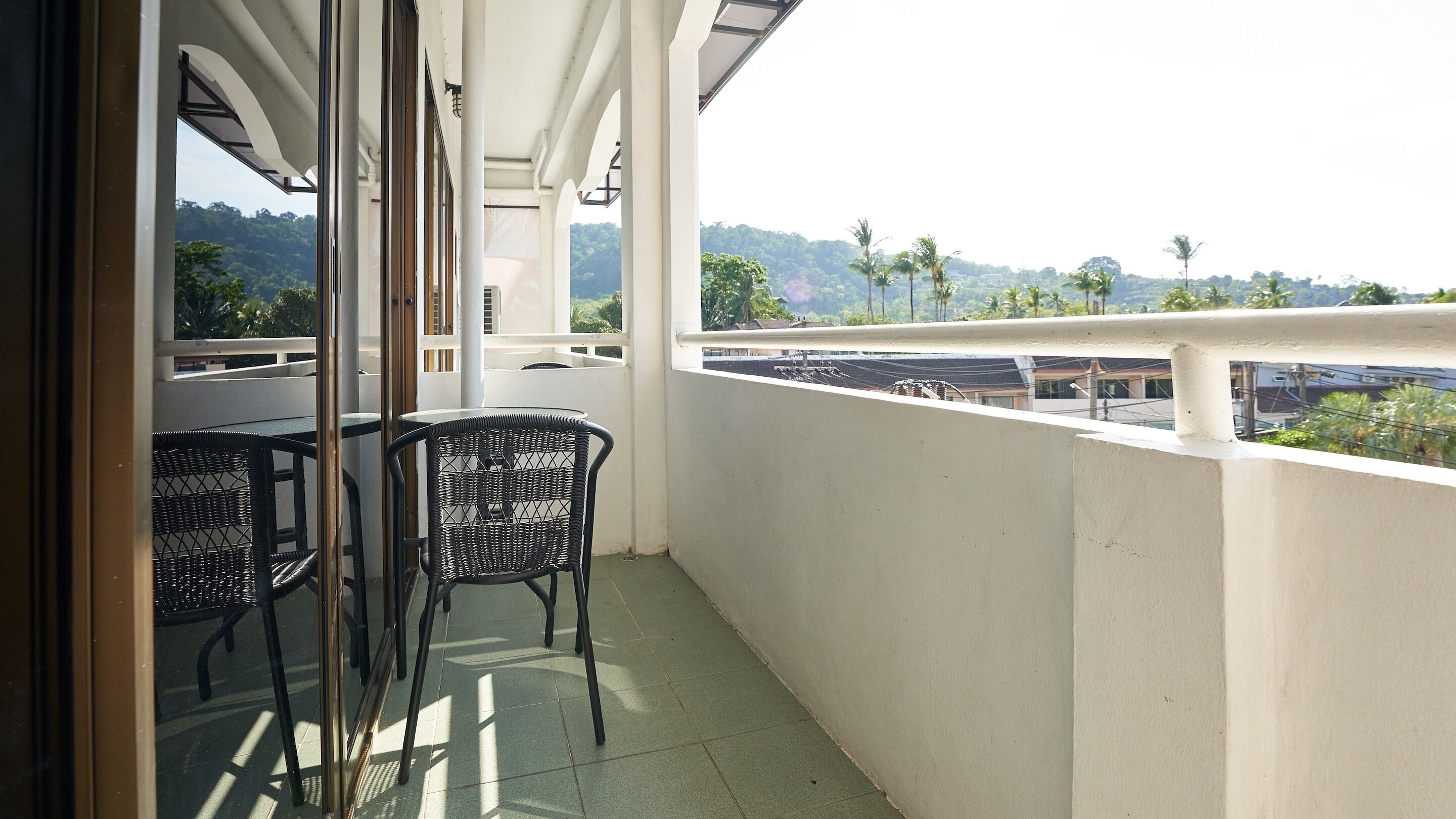A balcony with a glass door, two wicker chairs, and a view of a green landscape with trees and buildings.