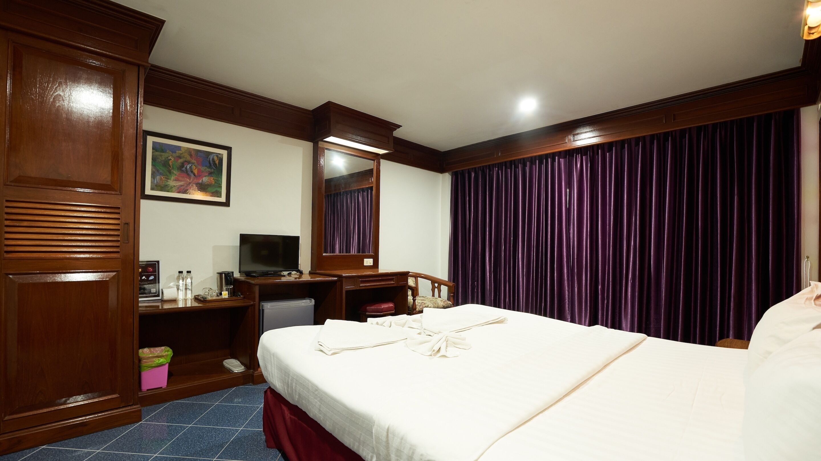 A hotel room with a bed, a desk with a television, a dresser, and a large window with purple curtains.