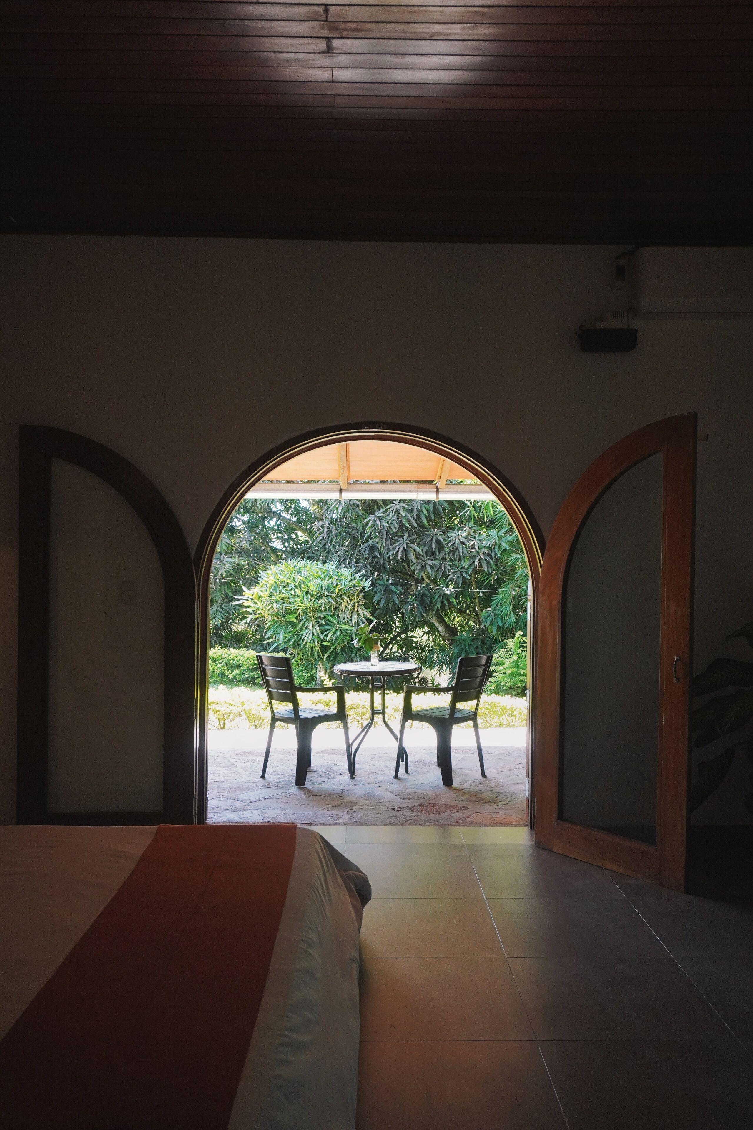 A room with a bed, an arched doorway leading to an outdoor seating area with chairs and a table, and a ceiling fan.
