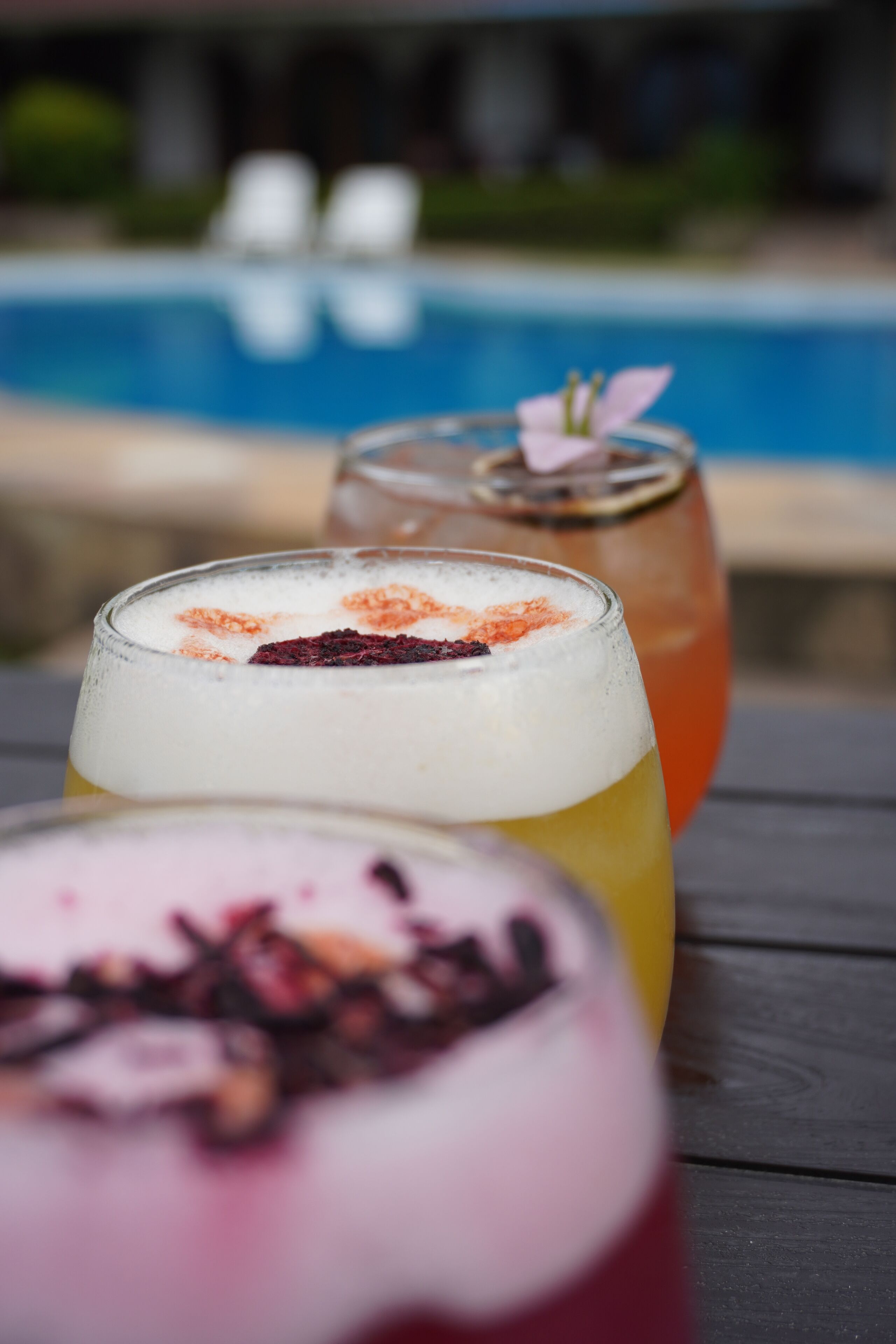 Três cocktails sobre uma mesa de madeira, com uma piscina ao fundo.