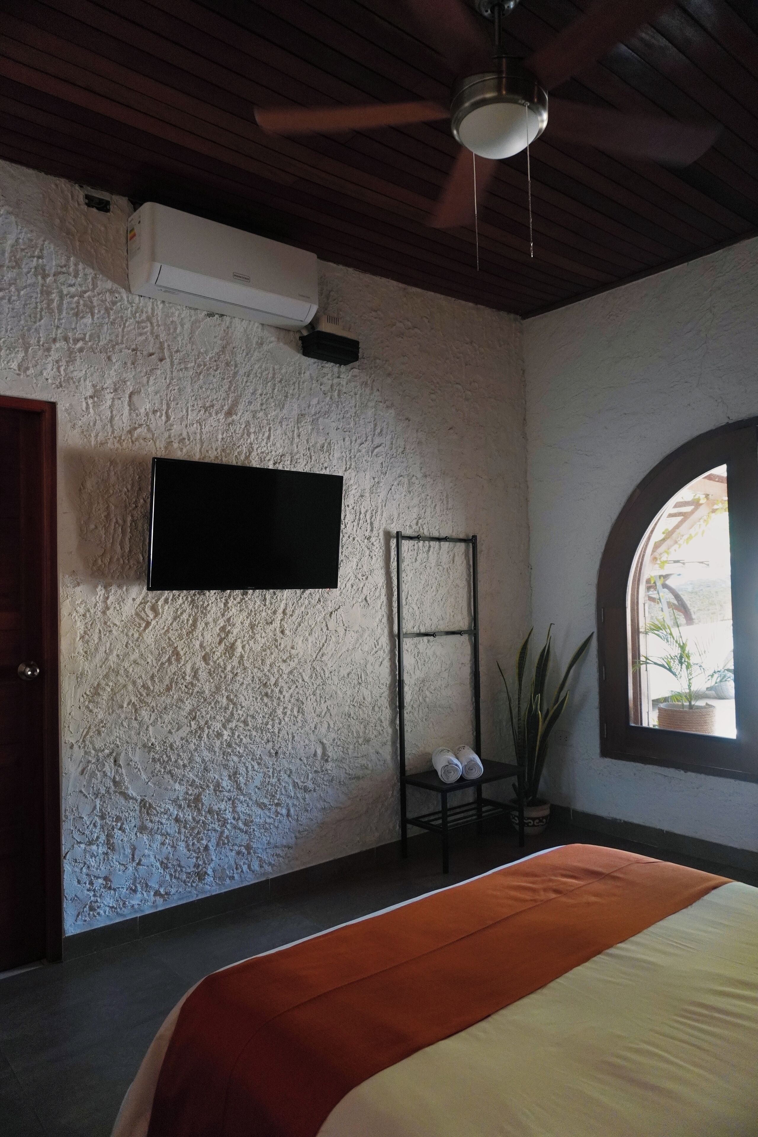 A bedroom with a bed, a ceiling fan, a mounted TV, a window with a view, and a wall-mounted air conditioner.