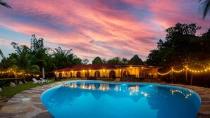 Una piscina con terraza de piedra, un porche cubierto con asientos y vistas a un edificio con ventanas arqueadas y luces colgantes.