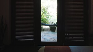 A room with a bed, a window with blinds, and a view of an outdoor area with plants. - APU SAMAY (La Banda de Shilcayo)