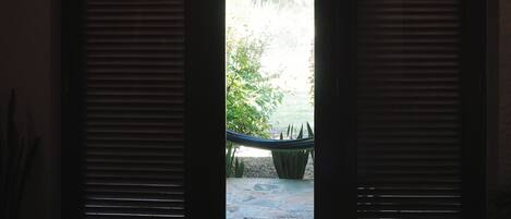 A room with a bed, a window with blinds, and a view of an outdoor area with plants.