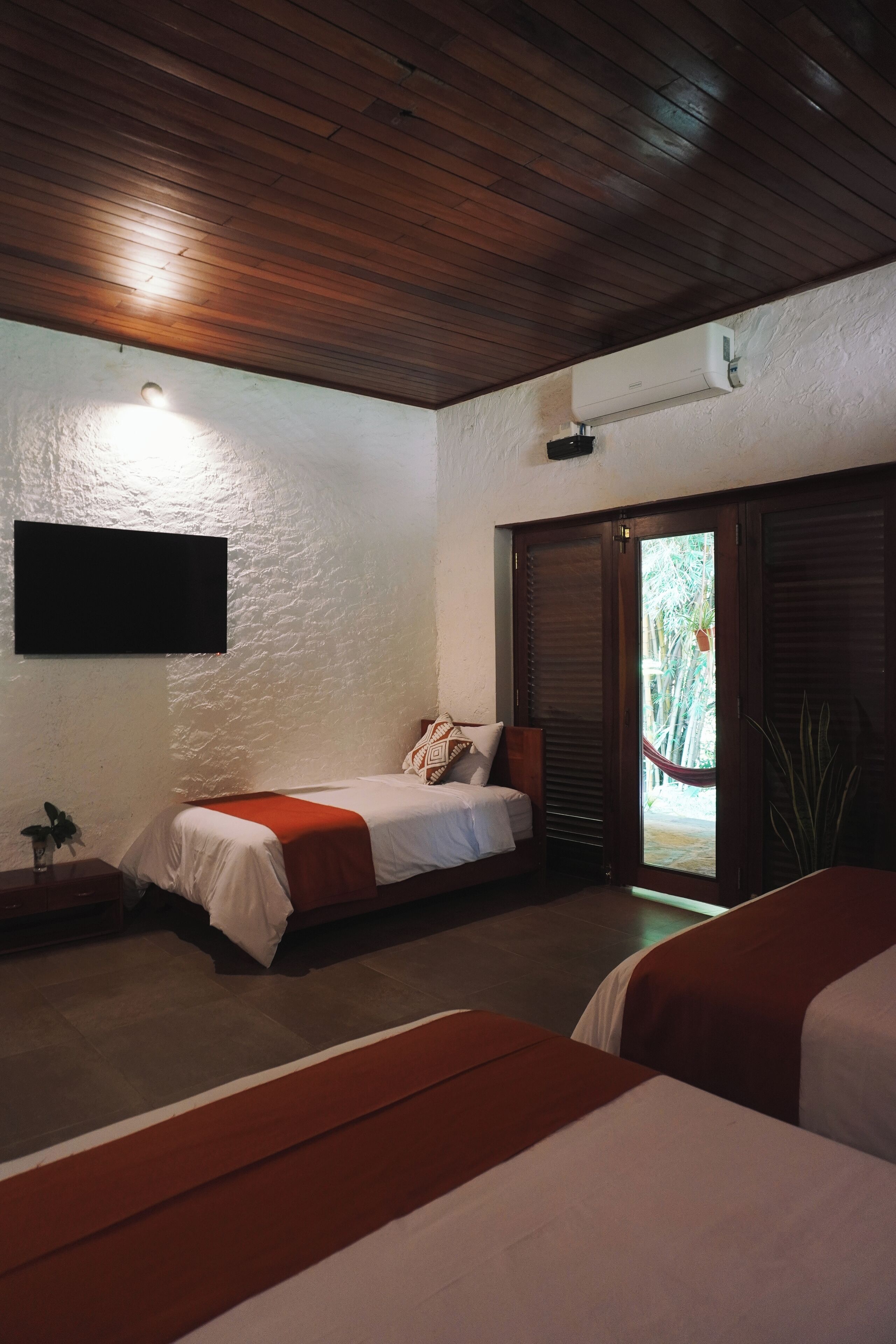 A bedroom with a wooden ceiling, a bed with a red and white bedspread, a bedside table with a plant, and a door leading to a balcony with a view of greenery.