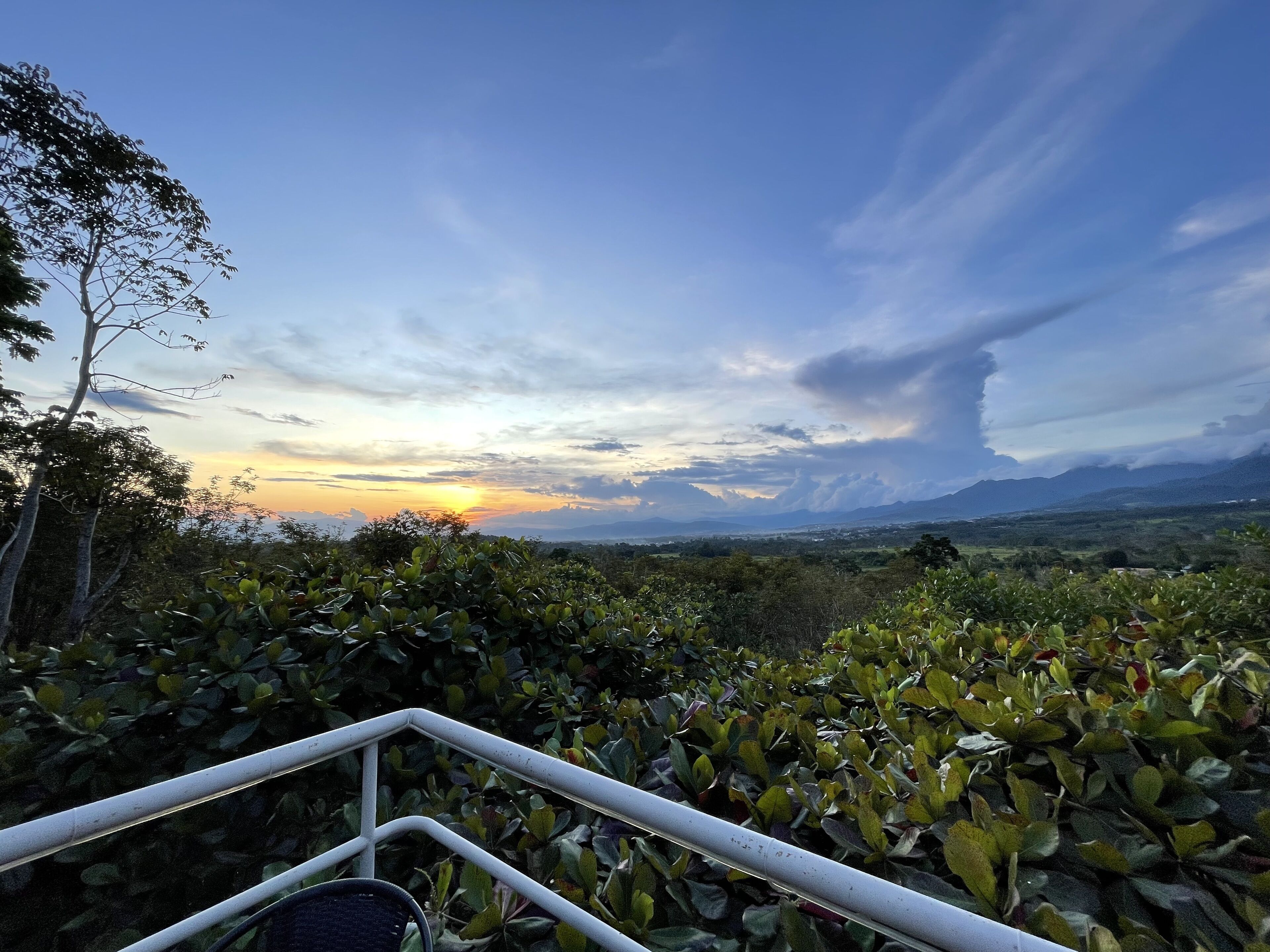 A scenic view from a balcony with a railing, overlooking a lush landscape and distant mountains during sunset.