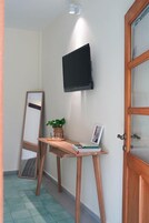 Una habitación con un escritorio de madera, un televisor de pantalla plana montado en la pared, un espejo grande apoyado contra la pared y una puerta de madera con paneles de vidrio.
