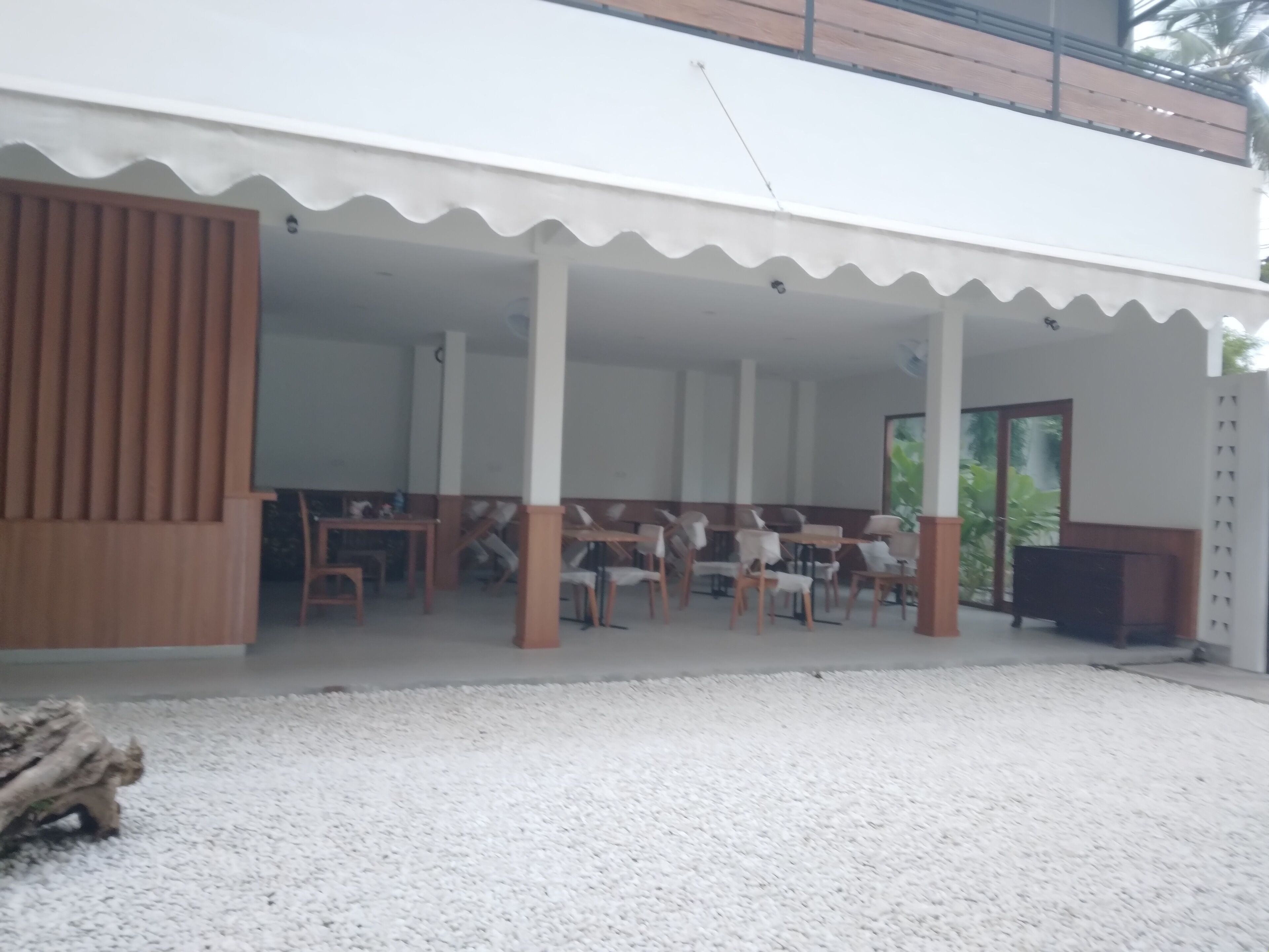 Un espacio de estar al aire libre cubierto, con mesas y sillas, techo blanco y vistas al verdor exterior.