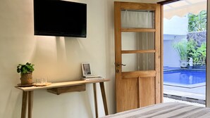 Una camera da letto con un letto, una TV a parete, una scrivania in legno con una pianta e una porta che dà su una zona piscina.