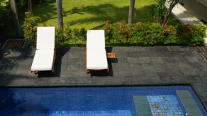 Una piscina con due lettini prendisole e un tavolino, circondata da un giardino con palme e arbusti.
