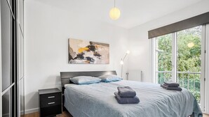 A modern bedroom with a large bed, a nightstand, a painting on the wall, and a window with a view of greenery. - Apartment - Sleeps 4 - Harbour Views - Parking (Aalborg)
