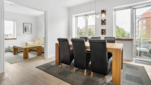 A modern dining area with a wooden table and black chairs, large windows with a view of buildings, and a sofa in the adjacent room. - Apartment - Sleeps 4 - Harbour Views - Parking (Aalborg)