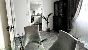 A modern living room with a glass dining table, two grey armchairs, a black cabinet, and a kitchen visible through an open door. - Remarkable 2-Bed Apartment in Castlebar (Castlebar)