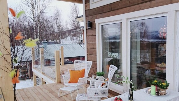 A wooden deck with white wicker chairs and a table set for dining, overlooking a snowy landscape.