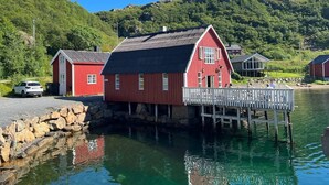 En rød træbygning med en anløbsbro, der strækker sig ud i rolige vand, omgivet af frodig vegetation og bakker.