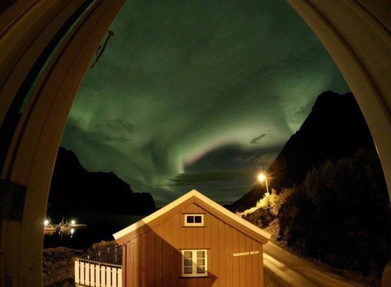 A wooden cabin with a sign reading "SANDO BLU" under a night sky with the aurora borealis.
