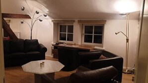 Un salon chaleureux avec un canapé en cuir noir, une petite table basse et une table d’appoint en bois. On y trouve une grande lampe sur pied et une fenêtre donnant sur l’extérieur.