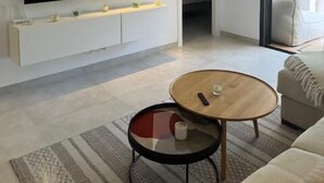 Un moderno salón con un televisor de pantalla plana grande montado en una pared blanca, una mesa redonda de madera, un sofá color beige y una alfombra estampada en el suelo.