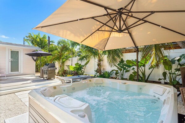 A backyard with a hot tub, patio furniture, and a grill under a large umbrella.
