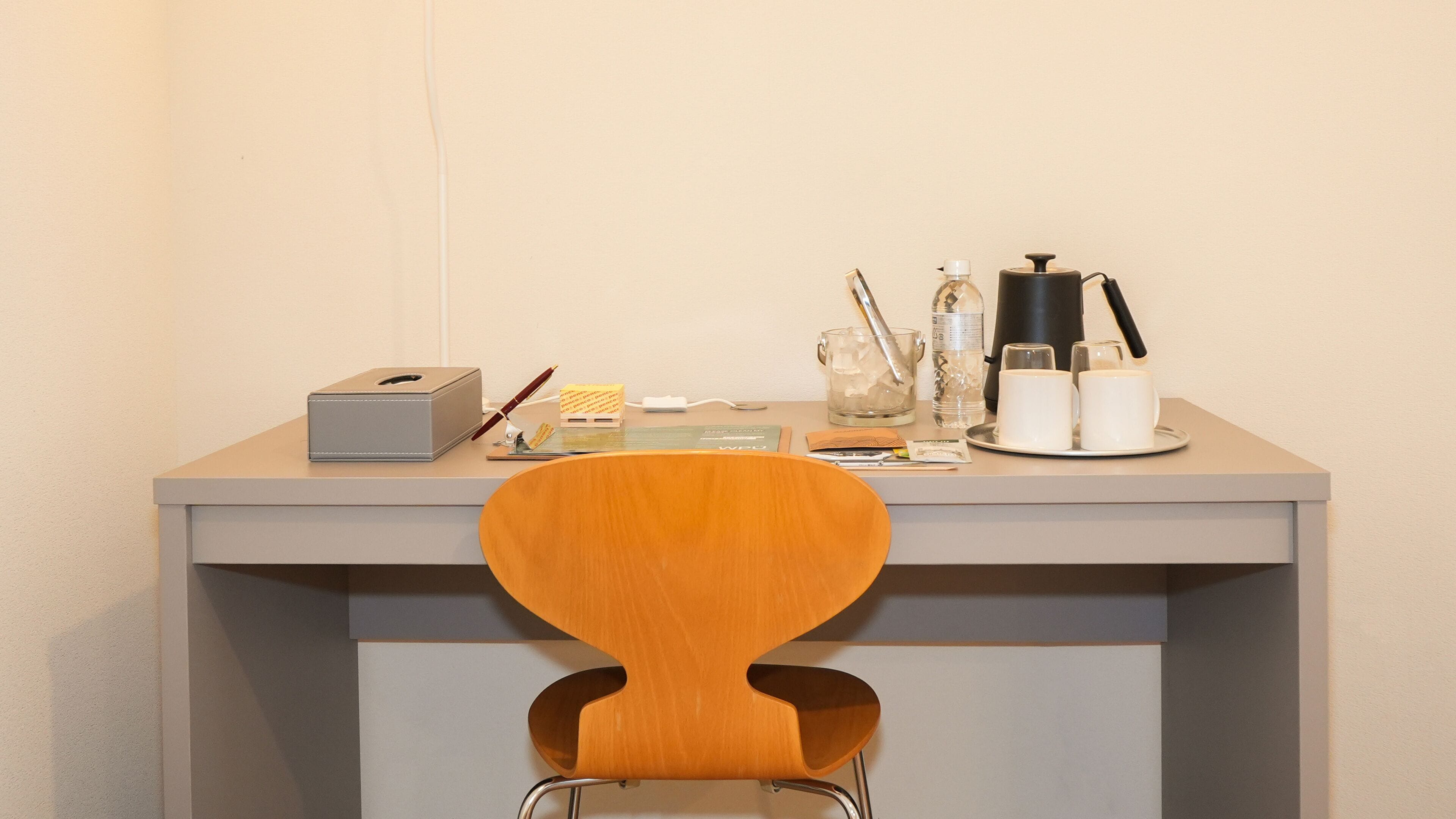 A desk with a wooden chair, a tissue box, a pen, a glass, a water bottle, a coffee maker, and a tray with two cups.