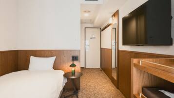 A hotel room with a bed, a bedside table with a lamp, a television mounted on the wall, and a carpeted floor.