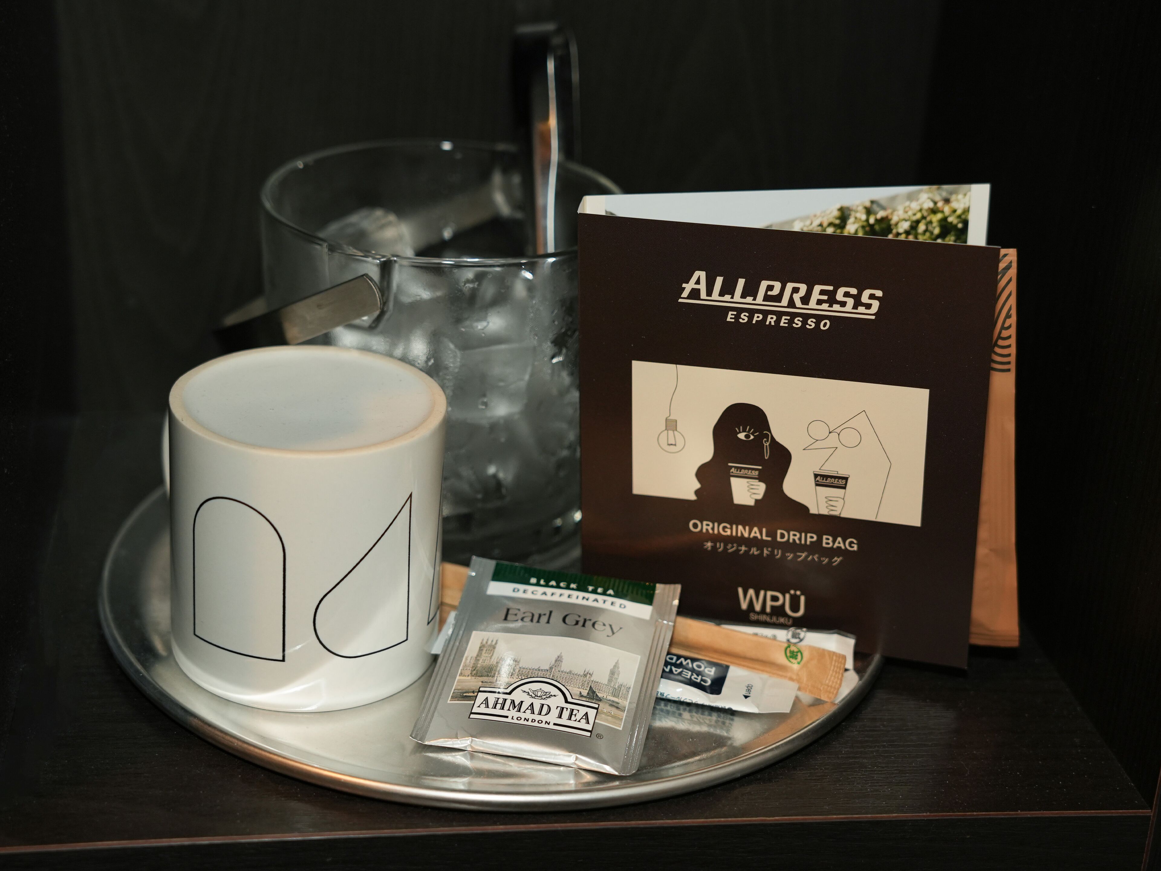 Una bandeja con una taza blanca, un vaso de agua, una bolsita de café de goteo y sobres de té.
