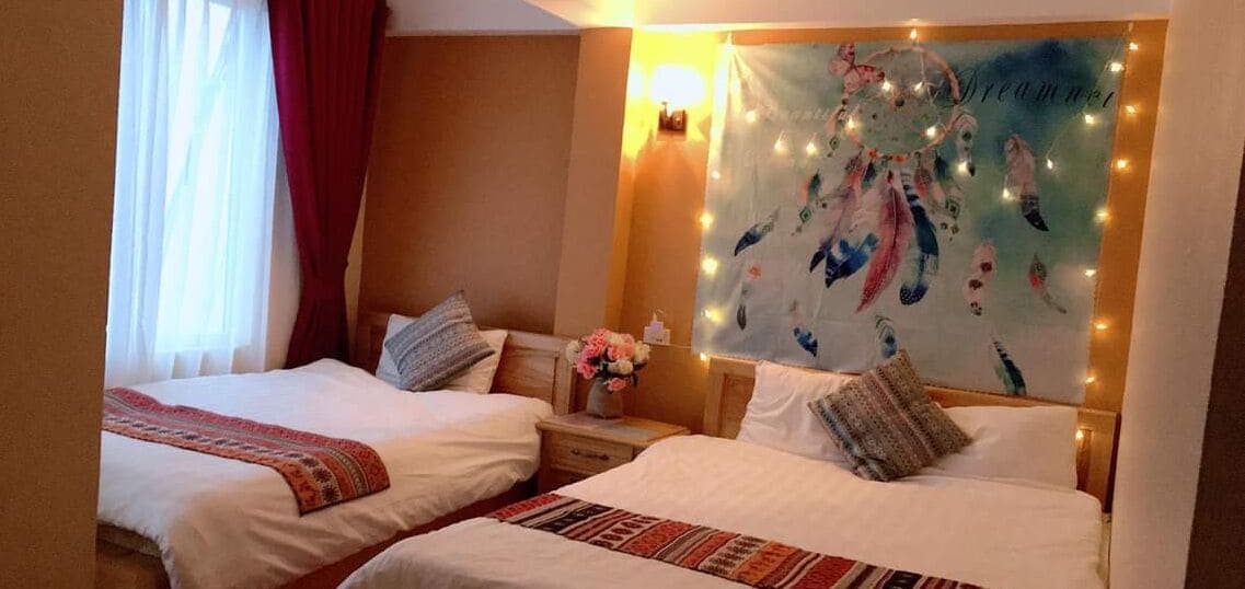 A hotel room with two beds, a nightstand with a vase of flowers, and a wall with a dreamcatcher and string lights.