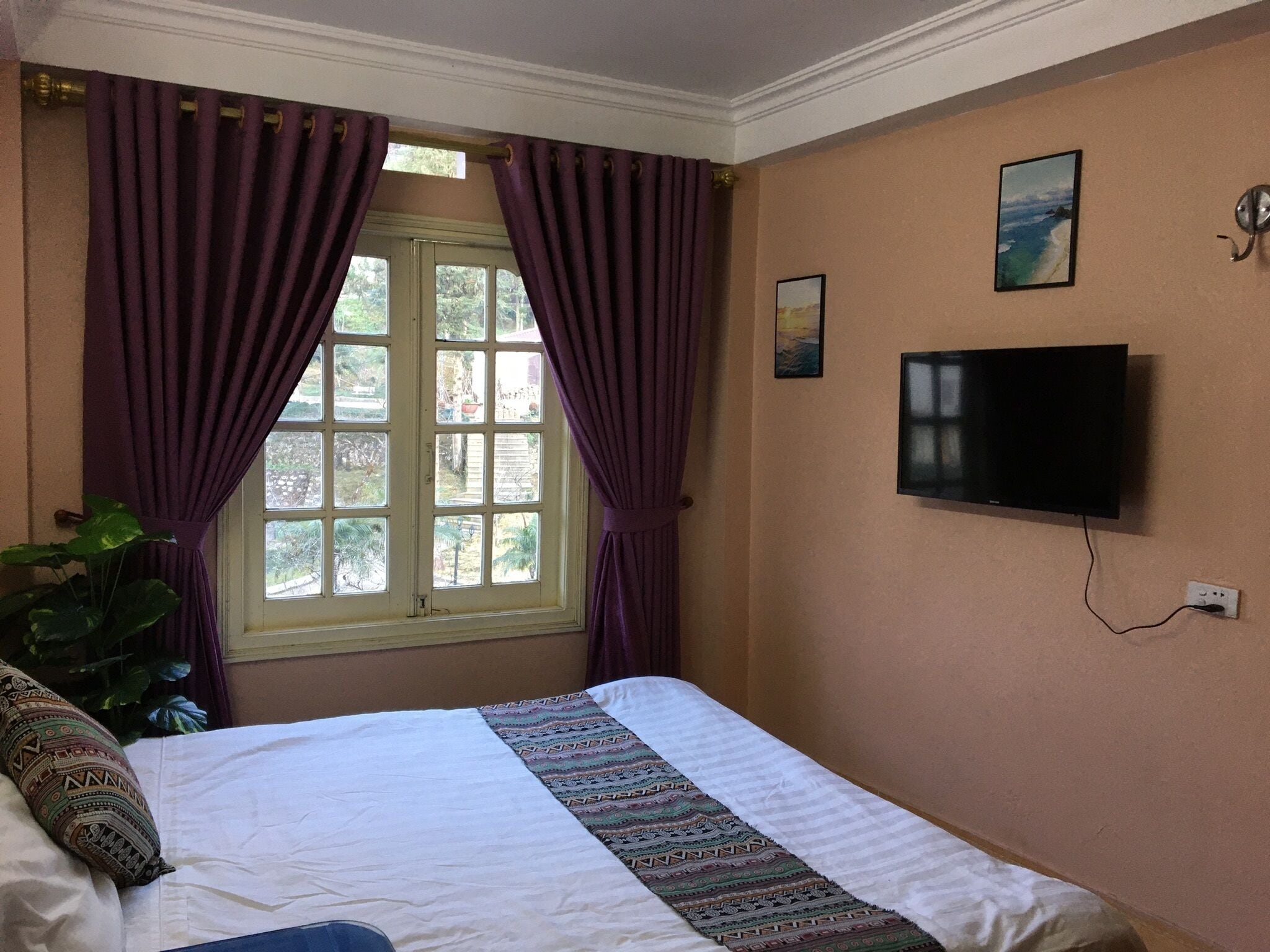 Un dormitorio con cama, una ventana con cortinas moradas, un televisor en la pared y un cuadro pequeño.