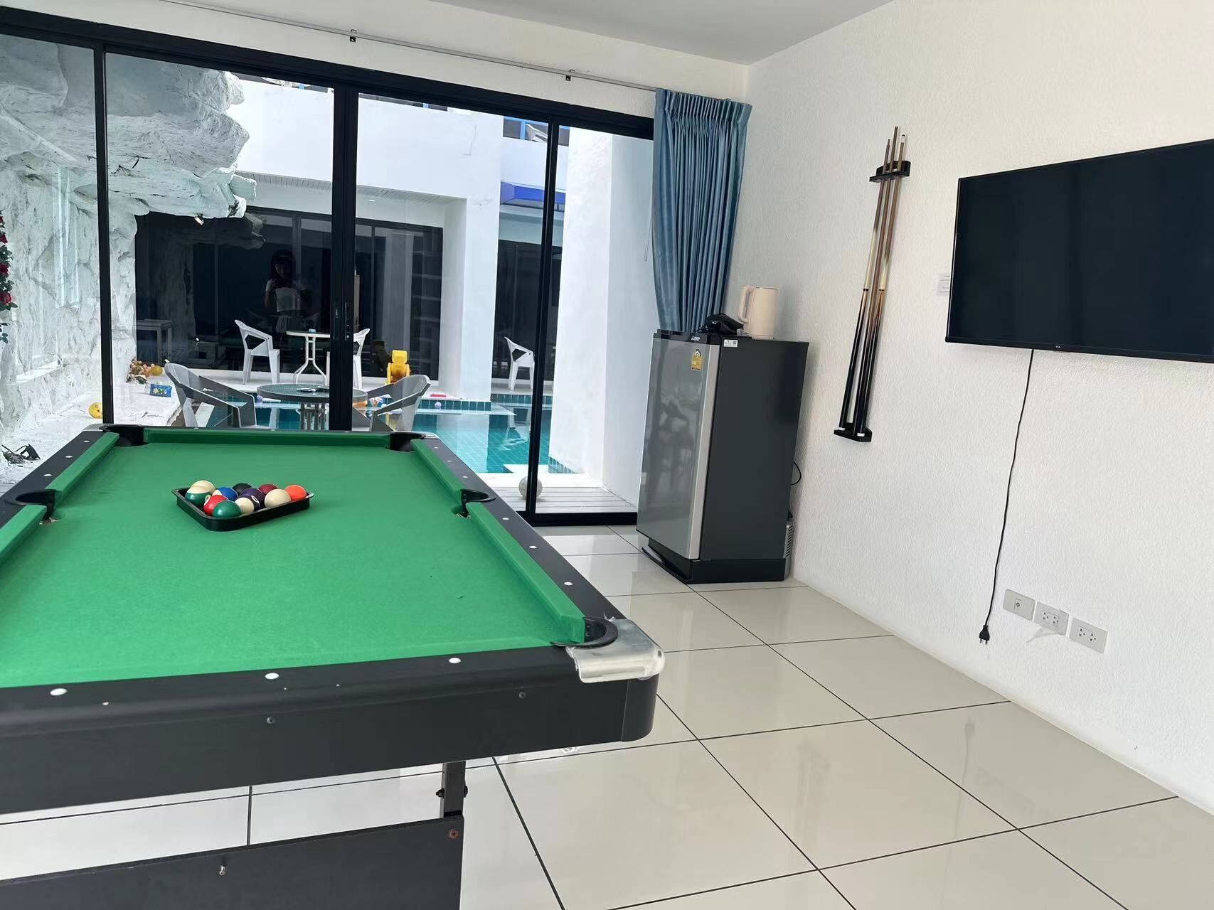 A pool table with balls, a refrigerator, and a television in a room with a view of a pool and outdoor seating.