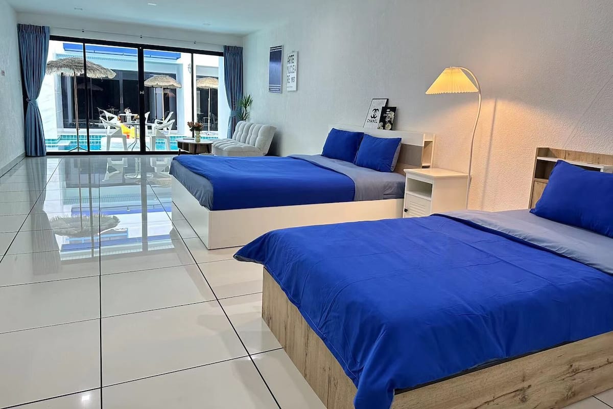 Ein modernes Hotelzimmer mit zwei Betten, einem kleinen Schreibtisch, einer Lampe und Blick auf einen Poolbereich.