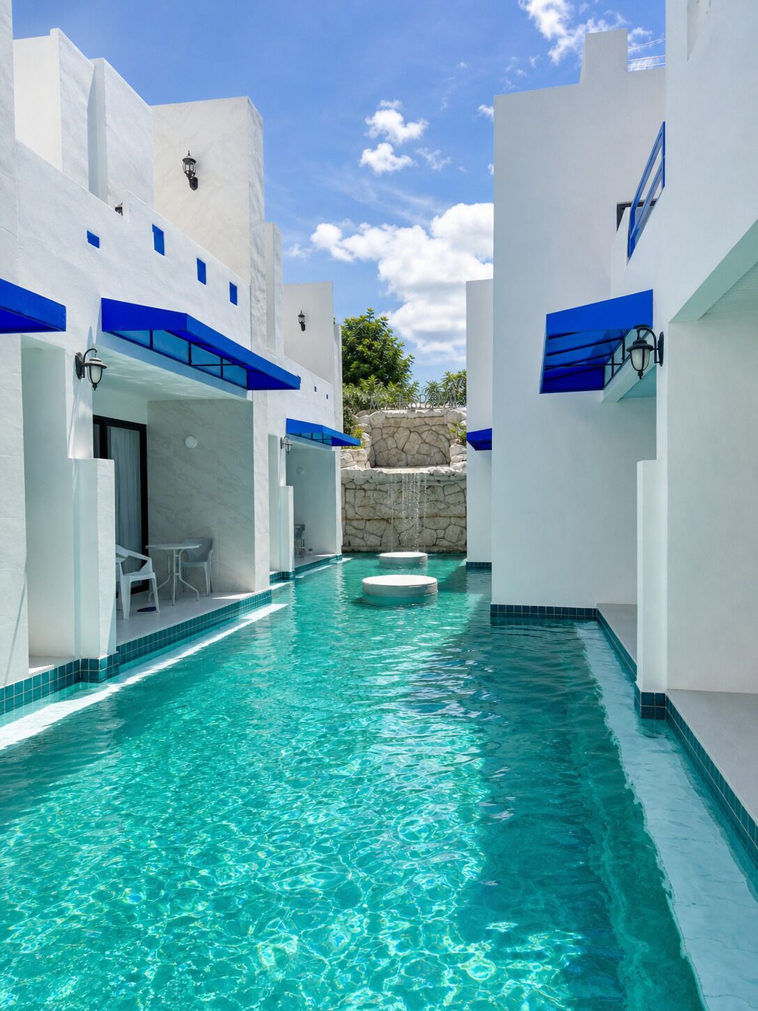 Una piscina con agua azul cristalina, edificios blancos con detalles azules y un muro de piedra al fondo.