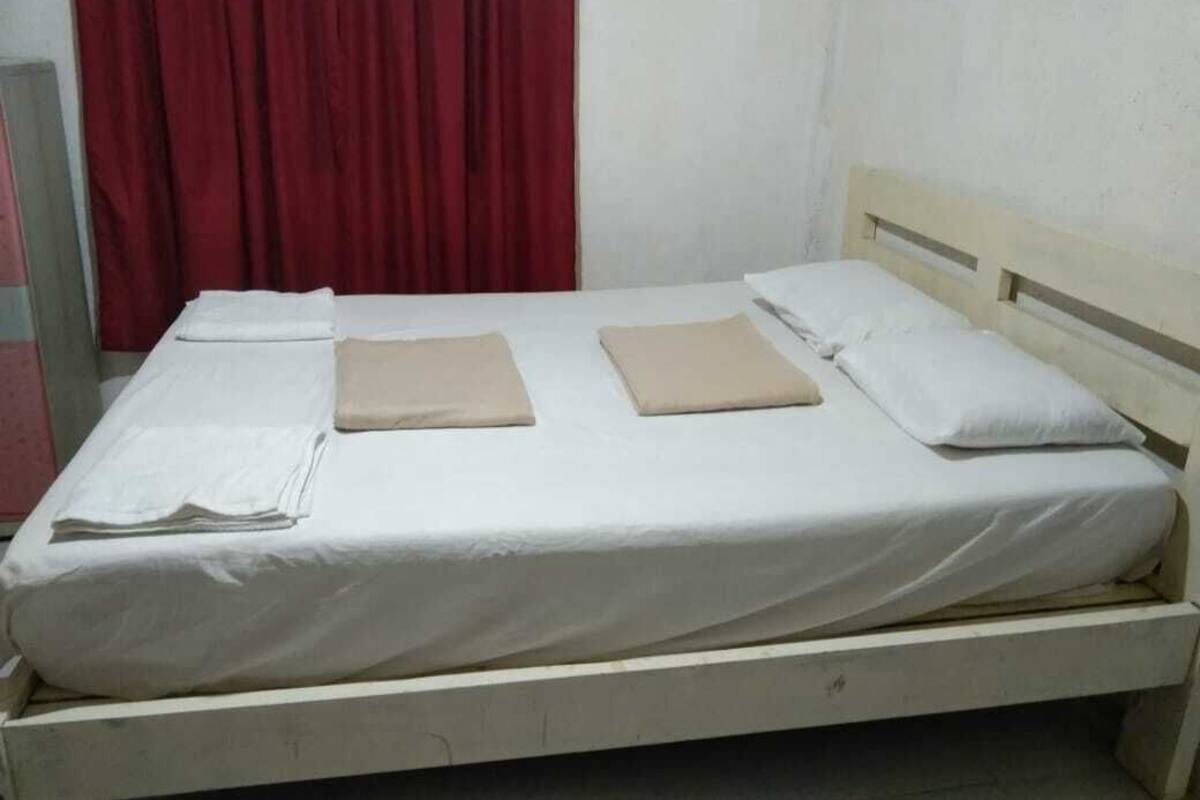 Un lit simple avec un matelas blanc et des oreillers beige, un rideau rouge et un mur uni.