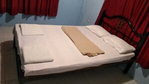 Uma cama de solteiro com colchão branco e manta bege, apoiada contra uma parede com cortinas vermelhas.