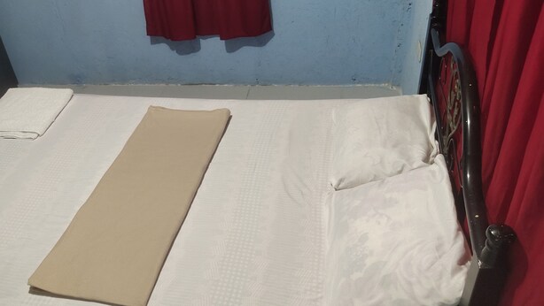 Uma cama de solteiro com colchão branco e manta bege. Há uma cortina vermelha no lado direito da cama.