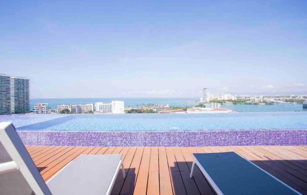 Una piscina infinita en la azotea con cubierta de madera y sillones de descanso, con vistas a un paisaje urbano y al mar.