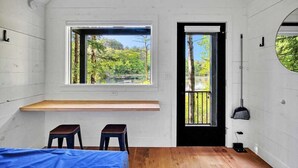 A room with a wooden bench, two stools, a bed, a window with a view of trees, and a door with a black frame. - Waterfront ADK Cabin and 2 Micro-cabins (Corinth)