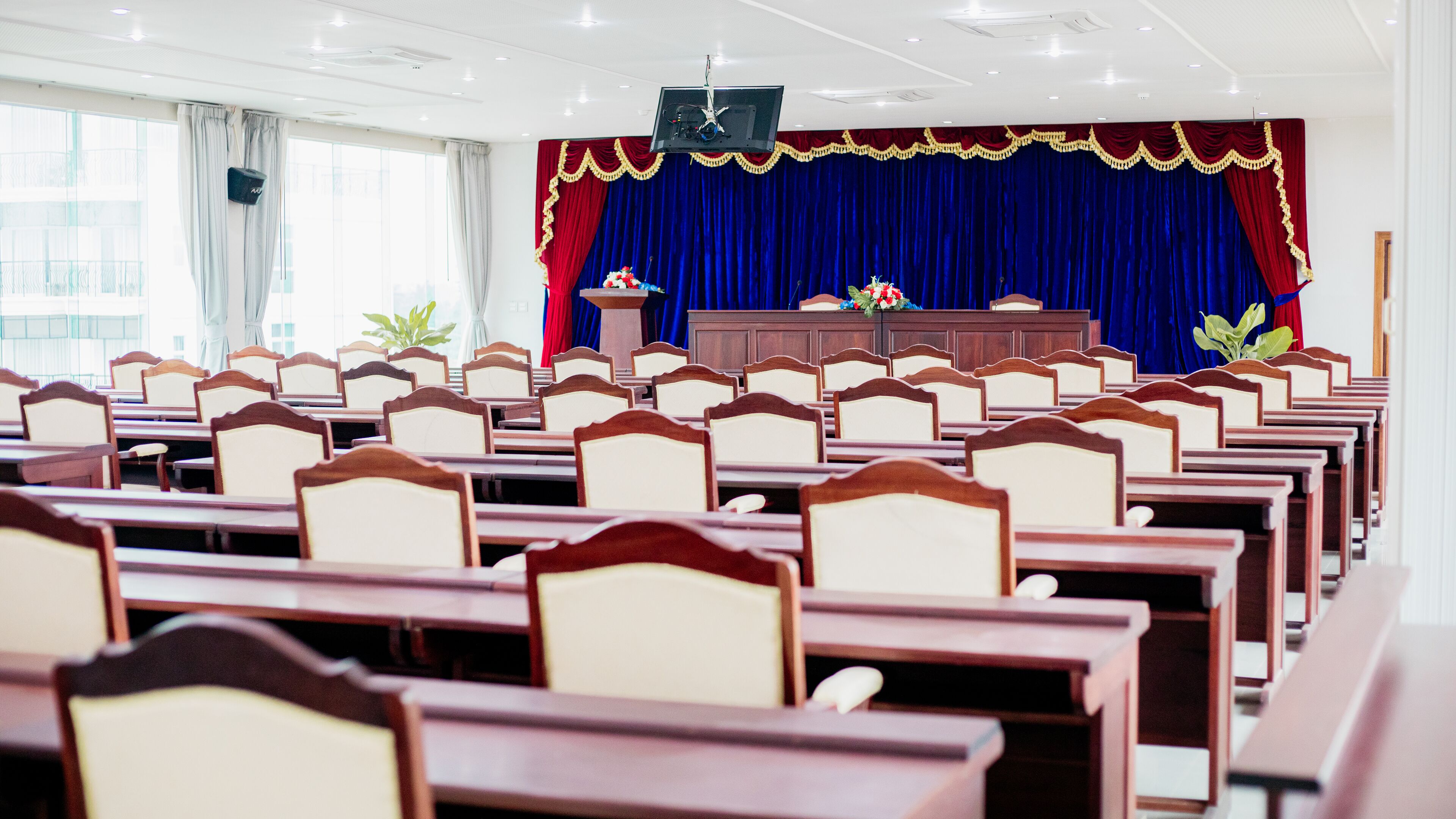 Une salle de conférence avec des rangées de bureaux et de chaises en bois, une scène avec un pupitre et un grand rideau bleu.