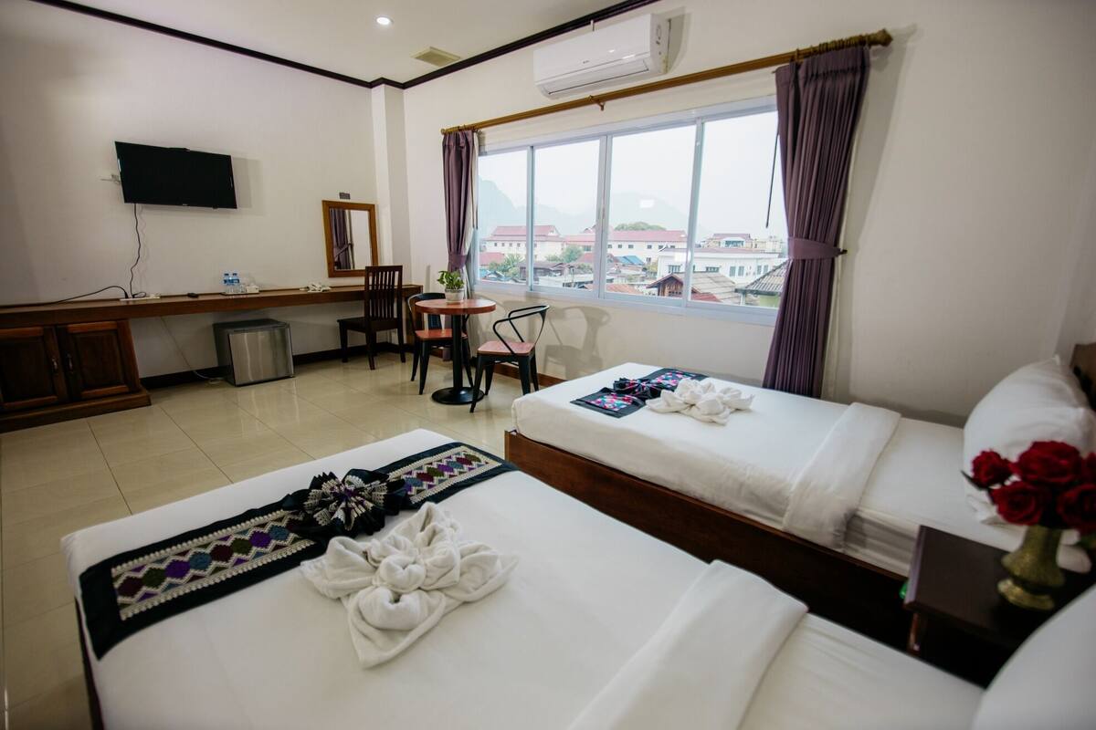 A hotel room with two beds, a desk, a chair, a TV, and a window with a view of buildings.