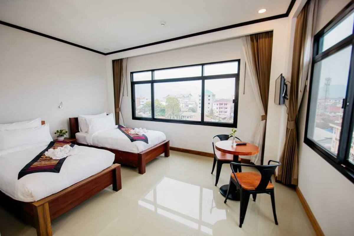 A hotel room with two single beds, a small round table with chairs, and a large window offering a city view.