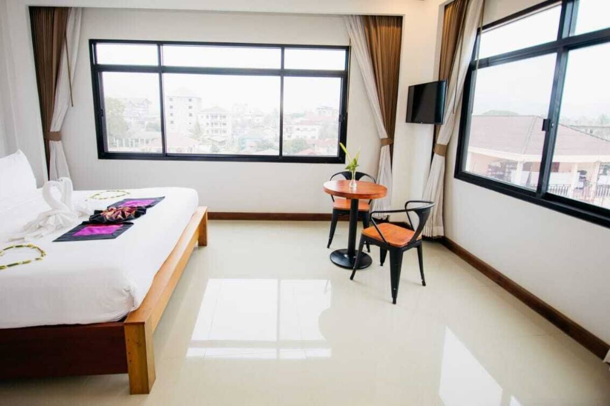 A hotel room with a bed, a small table with a chair, a TV, and large windows.