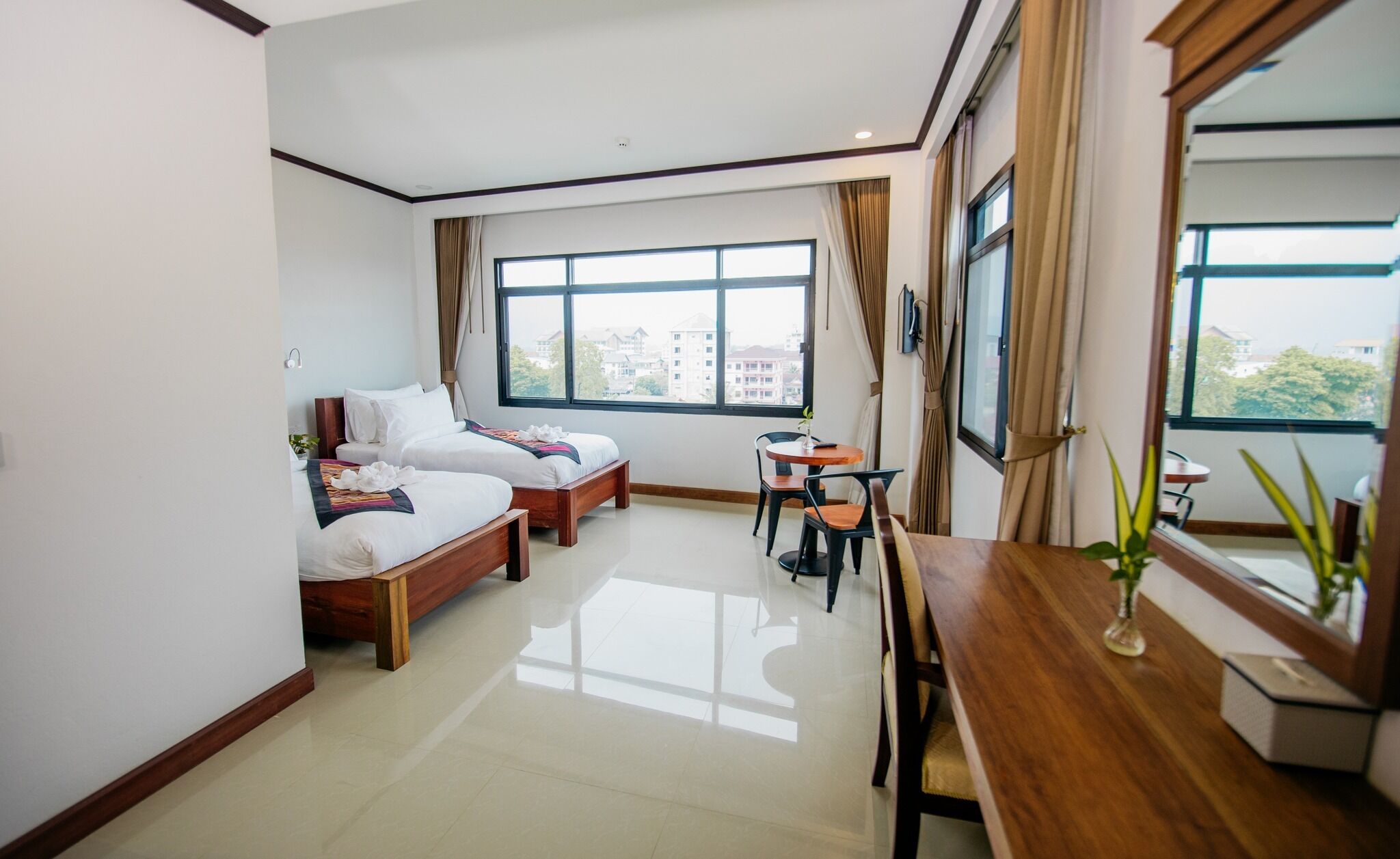 A hotel room with two beds, a dining table, and a large window with a city view.