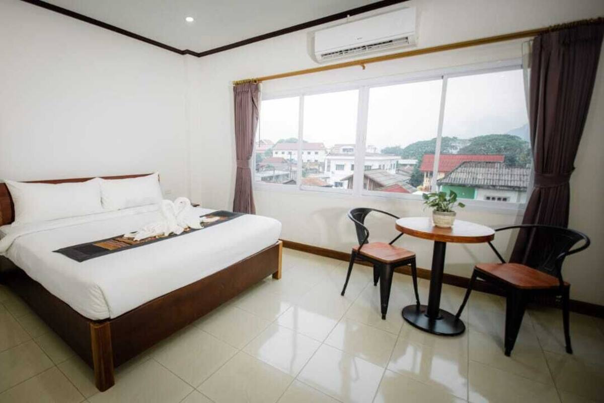 A hotel room with a large bed, a small round table with two chairs, a potted plant, and a window with a view of buildings.