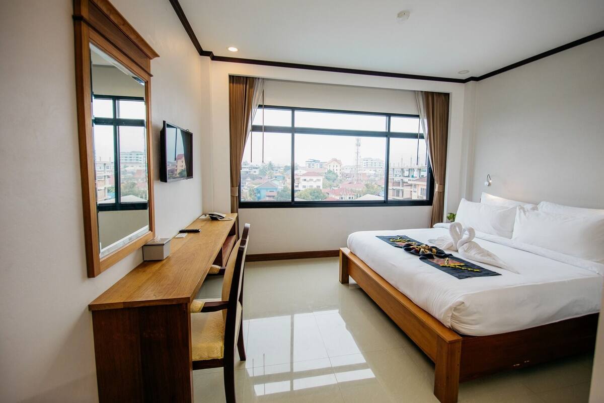 A hotel room with a bed, a desk, a chair, a TV, and large windows offering a city view.