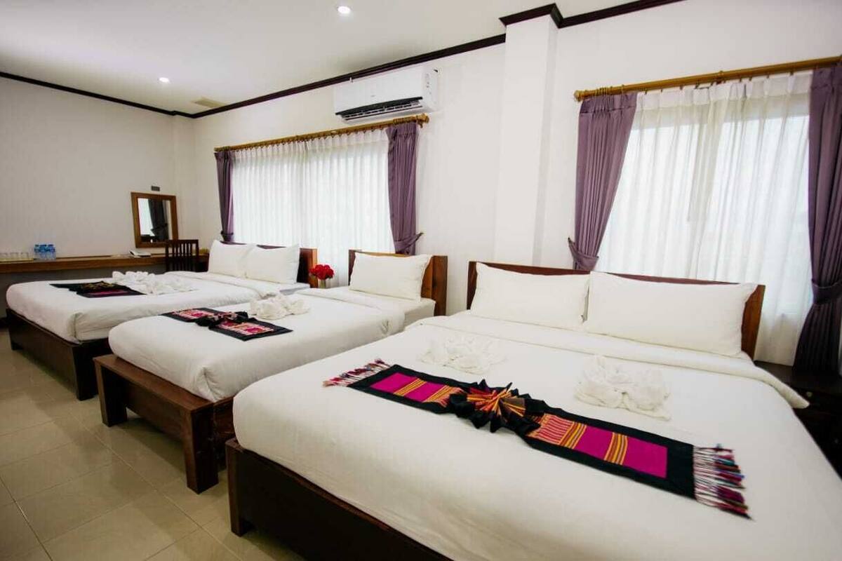 A hotel room with three single beds, each with white linens and decorative pillows. There is a large mirror on the wall and an air conditioning unit above it.