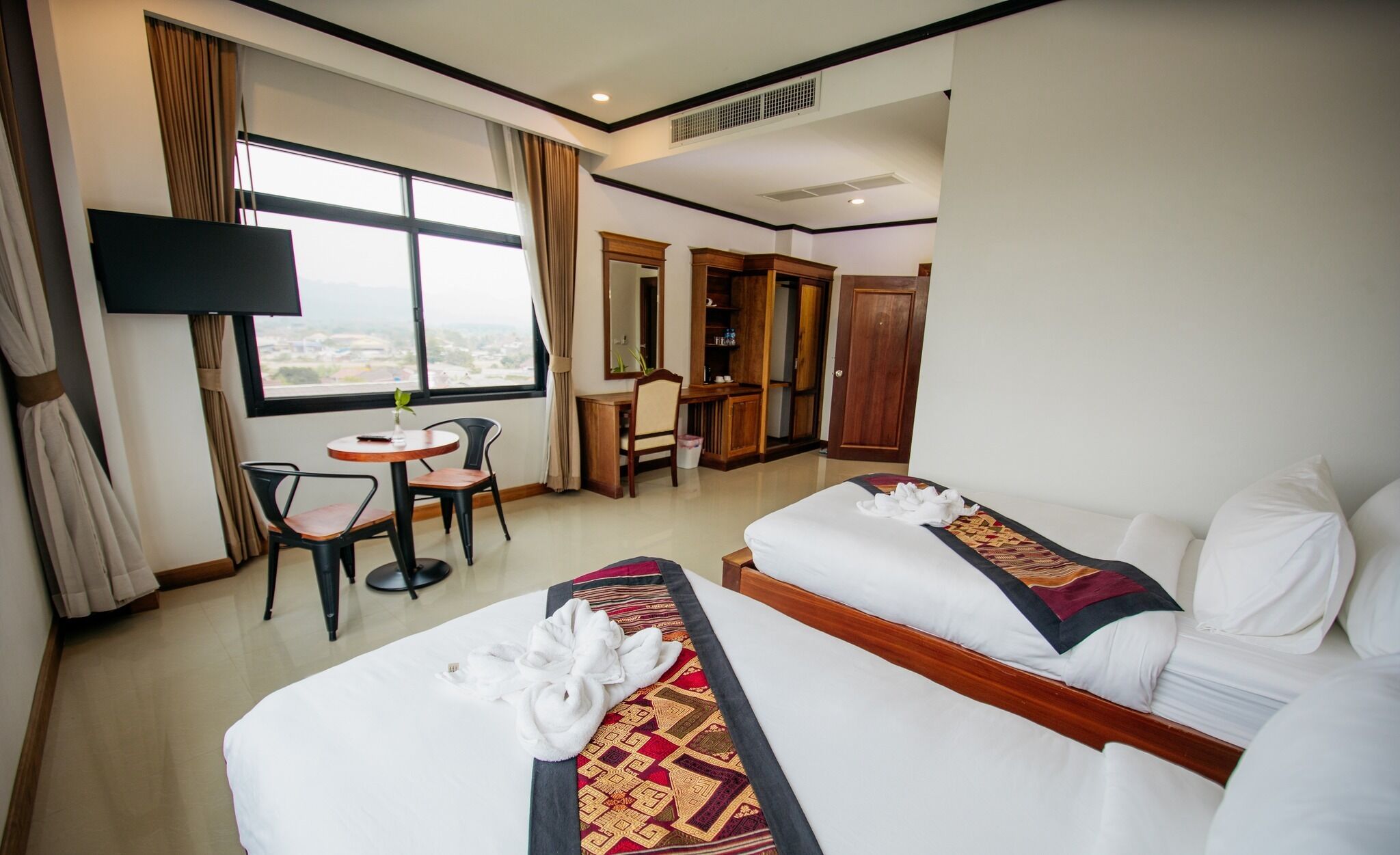 A hotel room with two beds, a desk, a chair, a TV, and a large window with curtains.