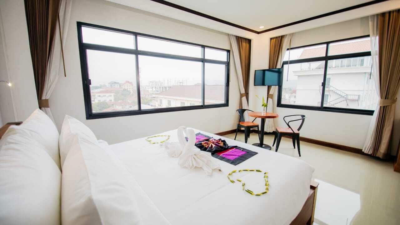 Ein Hotelzimmer mit einem großen Bett, einem kleinen Schreibtisch, einem Sessel und einem Fernseher. Vor den Fenstern hängen Vorhänge und es gibt einen Blick auf umliegende Gebäude.