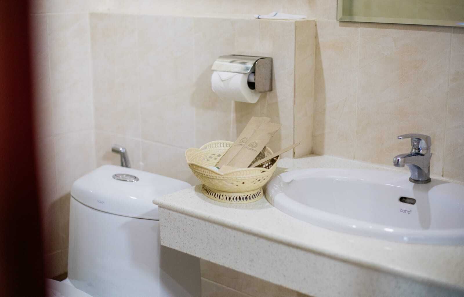 A bathroom with a toilet, a sink, a paper towel dispenser, and a basket with toiletries.