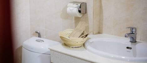 A bathroom with a toilet, a sink, a paper towel dispenser, and a basket with toiletries.